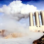 Female engineer recording data beside geothermal steam gushing from power station chimneys